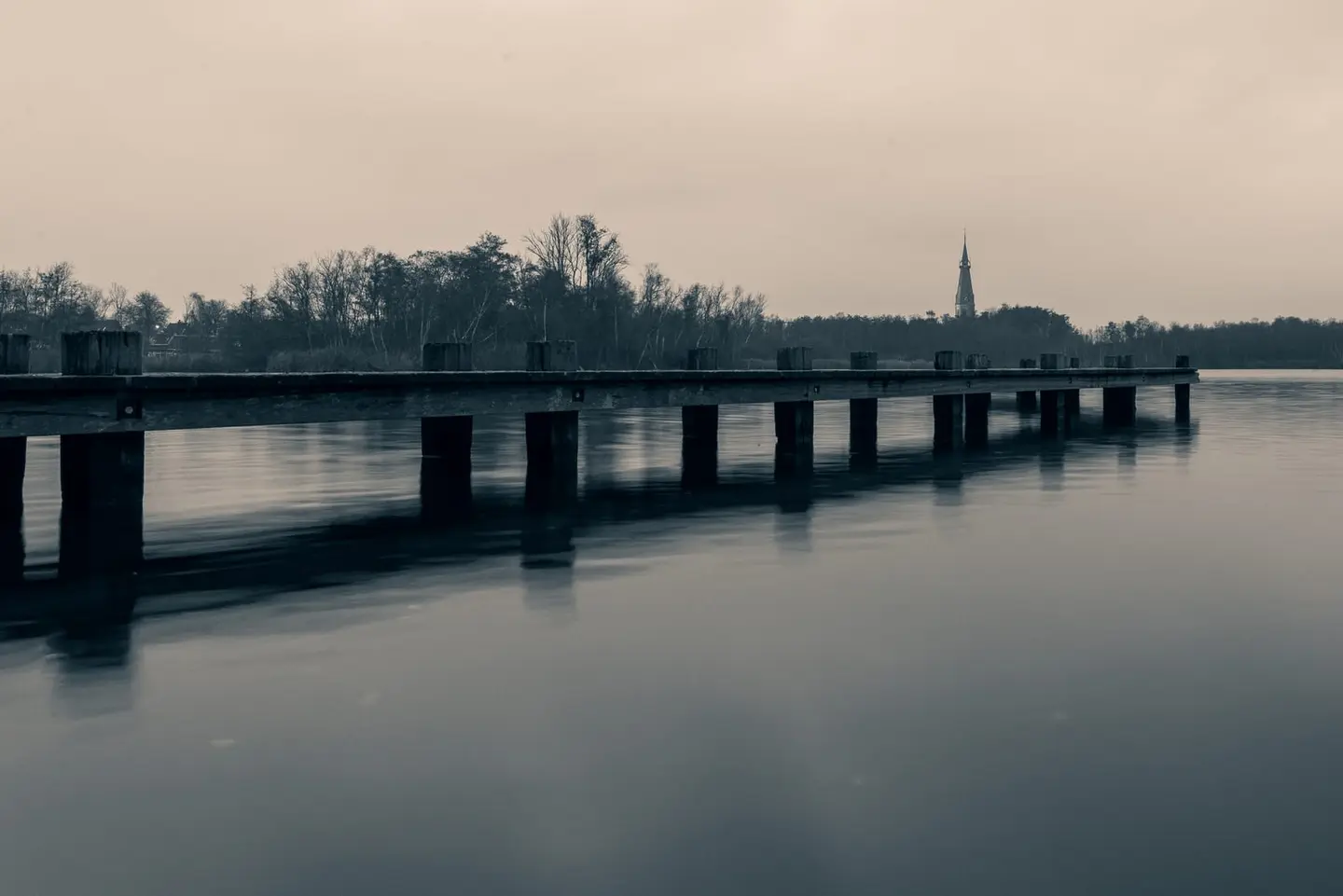 A weathered wooden jetty recedes toward the still surface of the Bosbaan lake in Amsterdam's urban forest, captured in the blue twilight of a November afternoon.